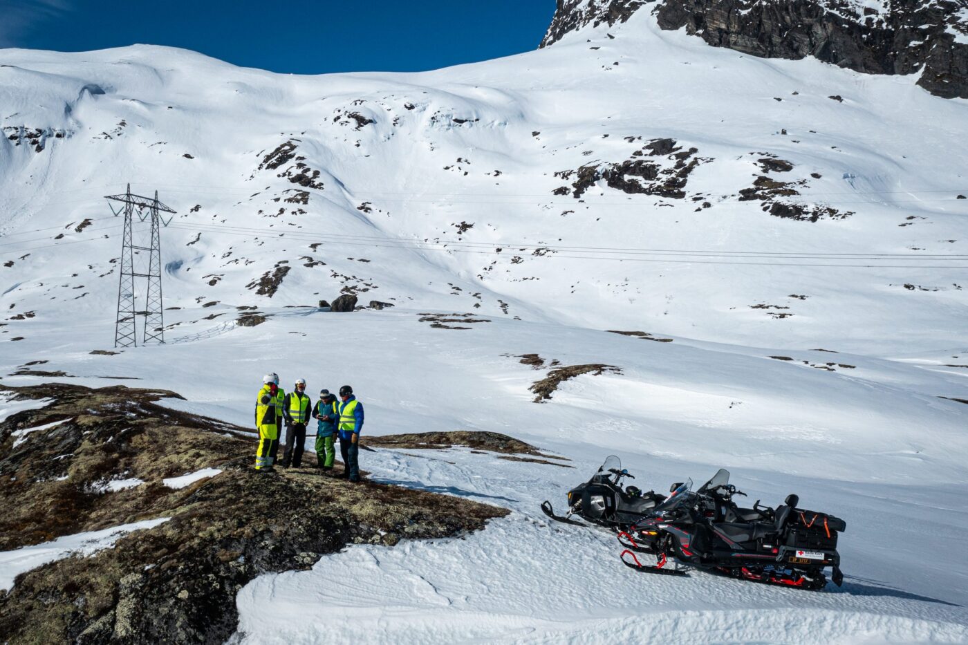 Fire personer i refleksvest står på en bergknaus i snødekt høyfjell ved siden av snøscootere