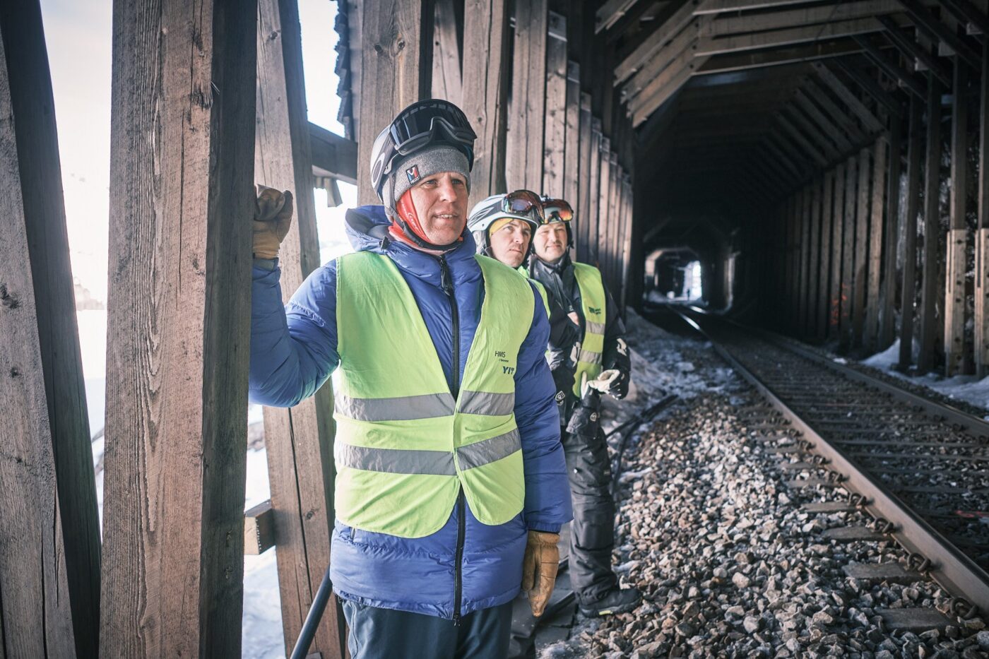 Person i refleksvest holder hånden mot en grov trestolpe i snøoverbygget, to andre står bak