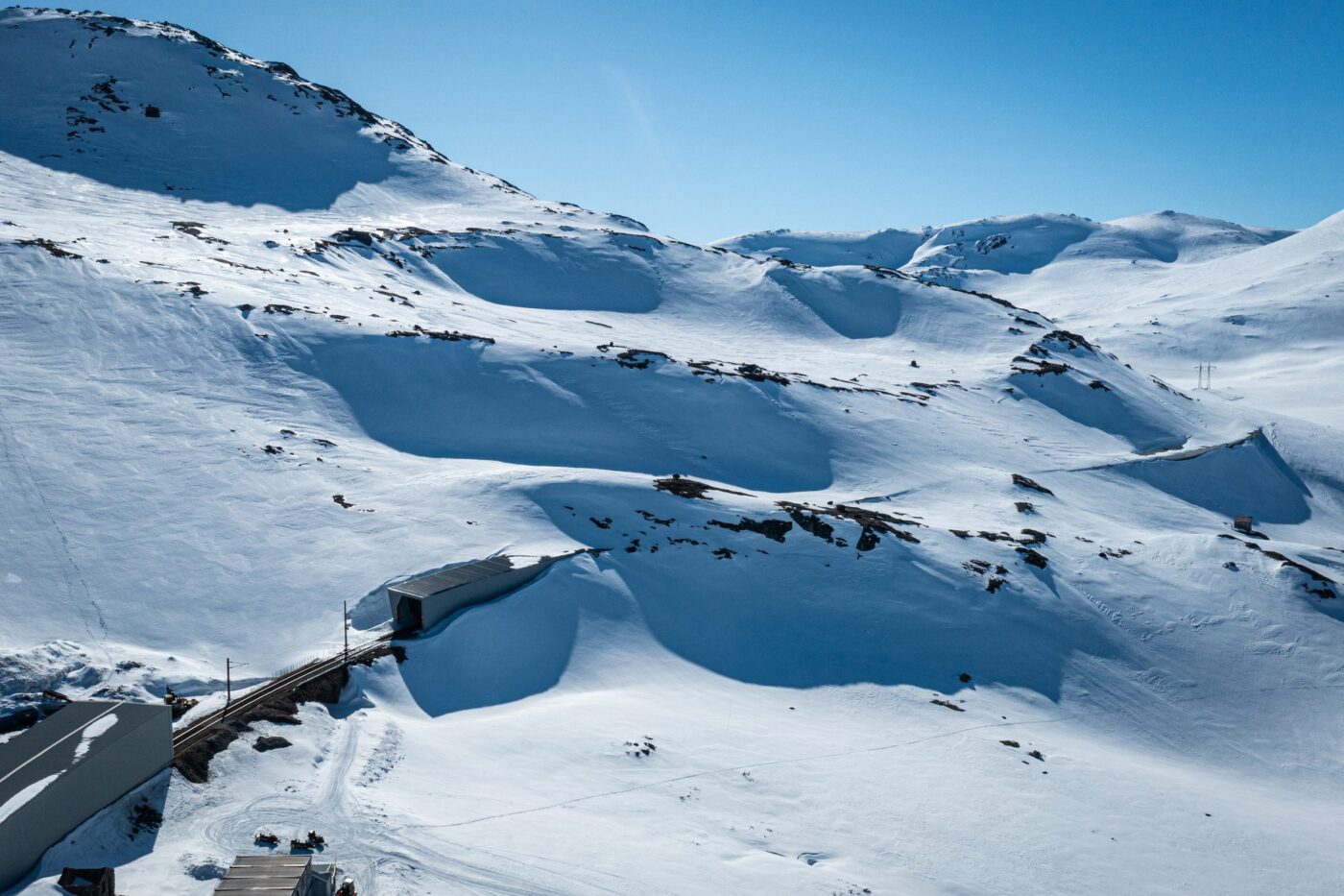 Luftfoto av snødekt høyfjell med jernbanelinje og snøoverbygg som en tynn strek i landskapet