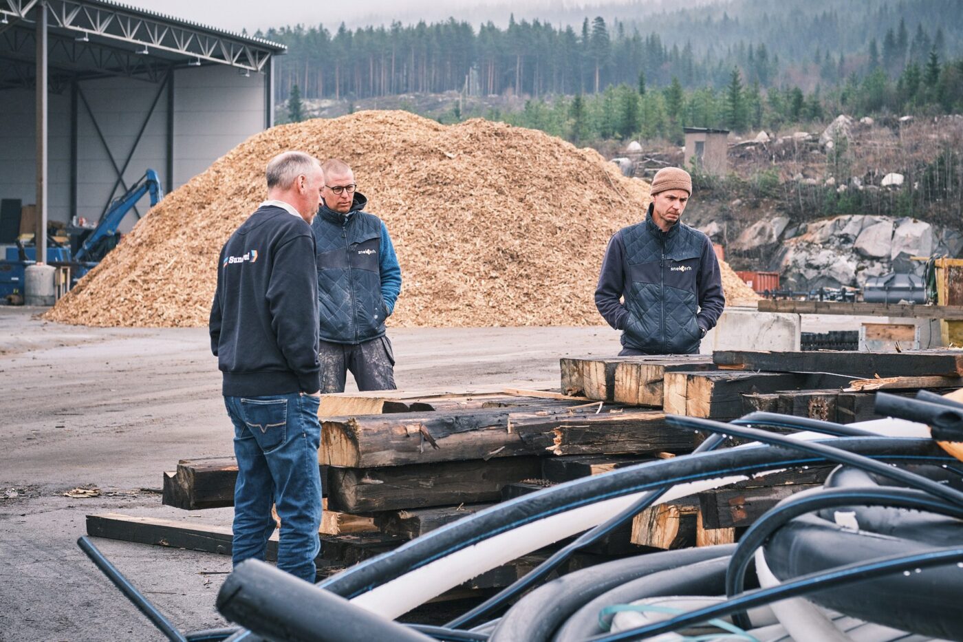 Tre menn i arbeidsklær står ved stabler av gammelt konstruksjonstømmer på et avfallsmottak