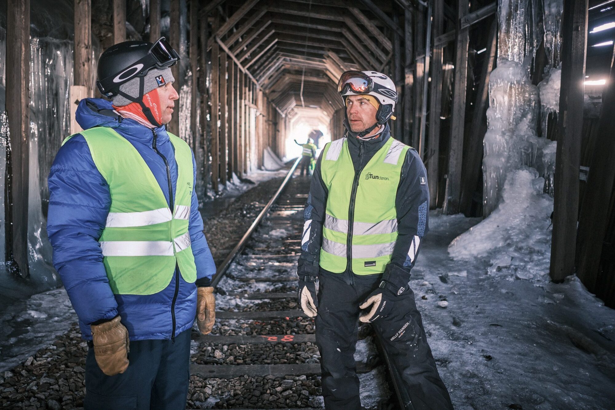 To personer i refleksvest står på jernbanesporet inne i et snøoverbygg, med is på veggene og lys i enden av tunnelen