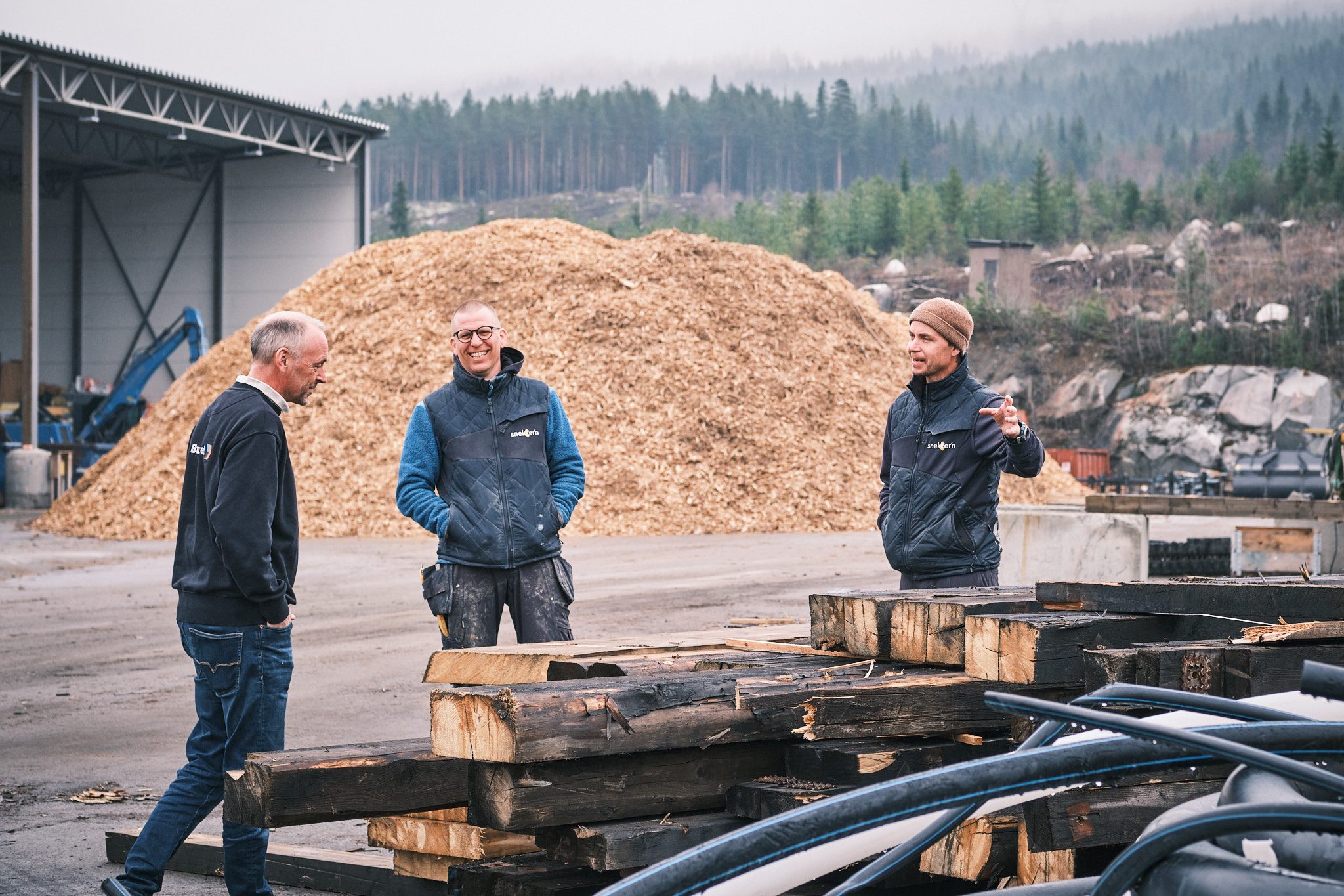 Tre menn i samtale foran en stor haug med flis på et avfallsmottak