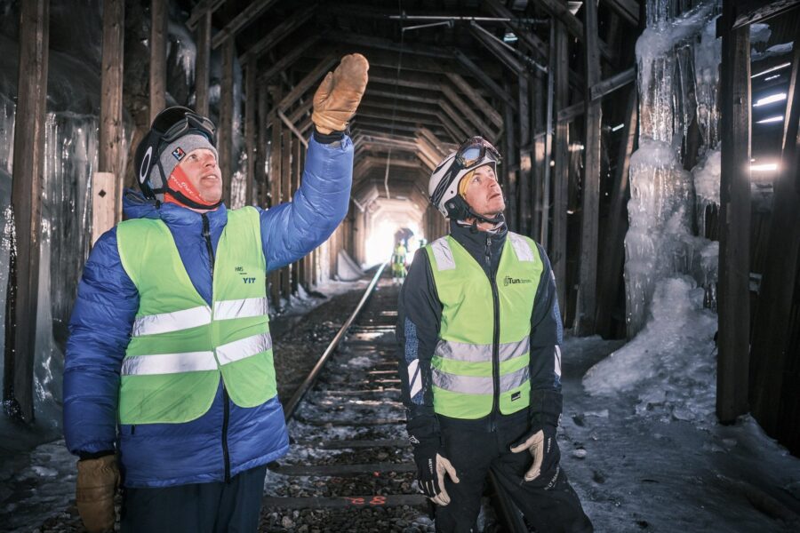 To menn i refleksvest inne i snøoverbygget, den ene peker opp mot takkonstruksjonen
