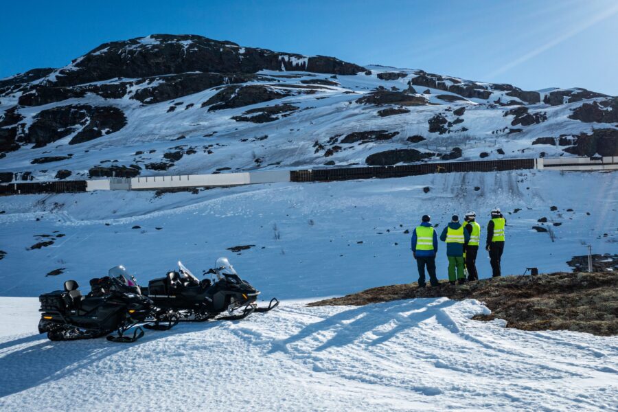 Fire personer i refleksvest på en bergknaus over snøoverbygget, med snøscootere