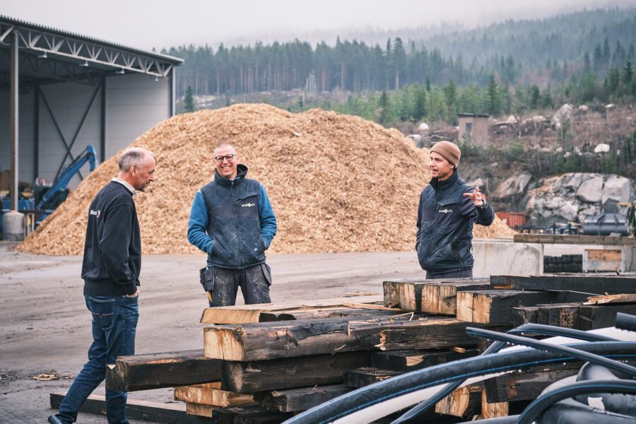 Tre menn i samtale foran en stor haug med flis på et avfallsmottak