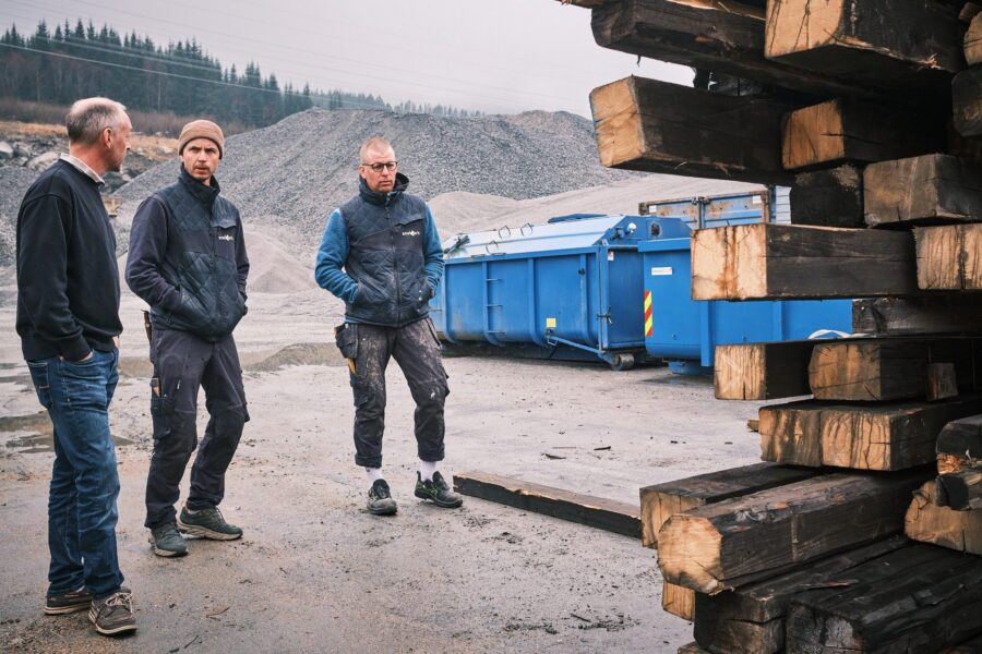 Tre menn ved en container med stabler av gammelt konstruksjonstømmer